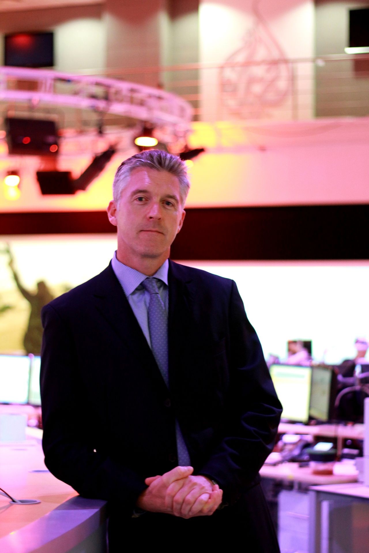 Man in a suit standing in a modern office with computers and screens in the background.