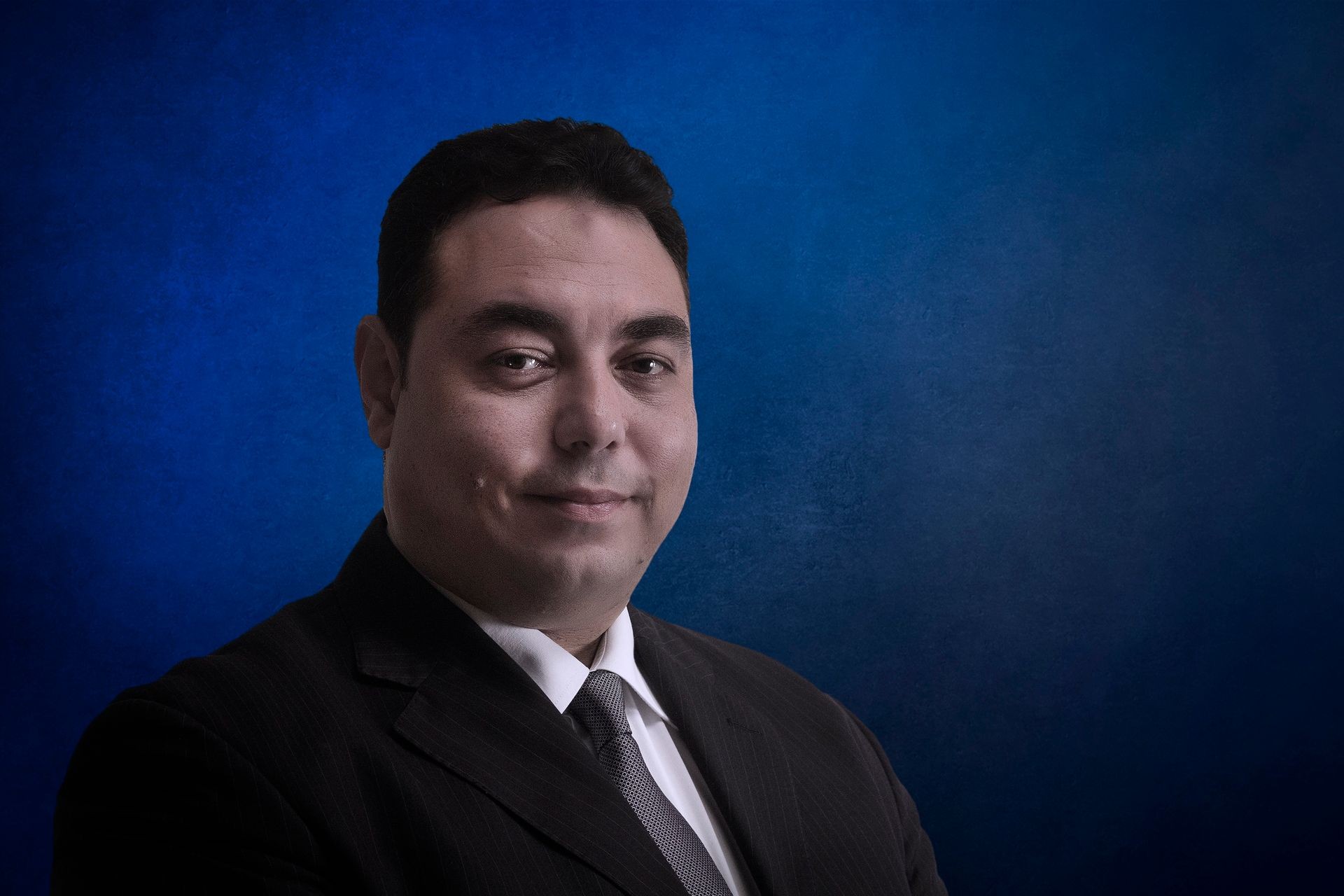 Person wearing a dark suit and tie in front of a blue background.